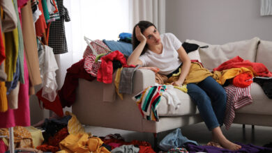 Upset young woman with lots of clothes on sofa in room. Fast fashion wardrobe