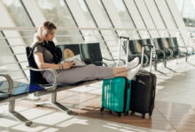 Passenger sitting on chair in modern light airport with outstretched legs on suitcases, reading magazine and waiting airplane arriving, flight delay. Young woman travel and relax on vacation.