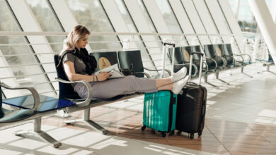 Passenger sitting on chair in modern light airport with outstretched legs on suitcases, reading magazine and waiting airplane arriving, flight delay. Young woman travel and relax on vacation.