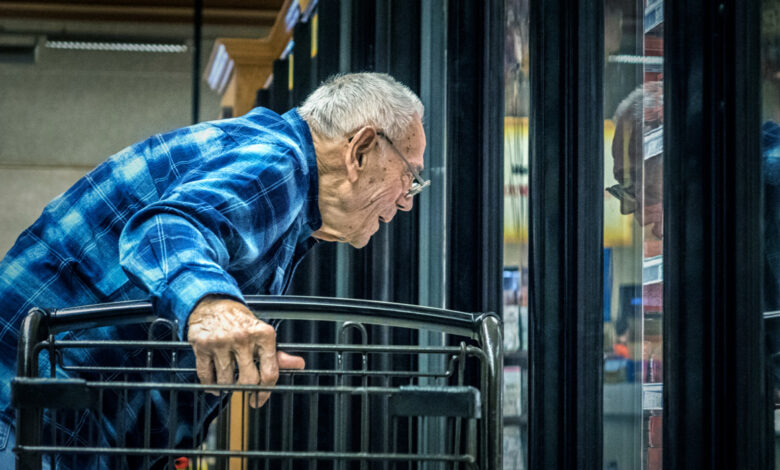 An indecisive, real person, elderly 96 year old senior adult man grocery shopper - supporting himself with his hand by holding onto his shopping cart - is leaning down in front of a supermarket grocery store refrigerated section cooler door looking at multiple potential frozen food dinner choices. His uncertain face is reflected in the refrigerator glass window. Image has some grain.