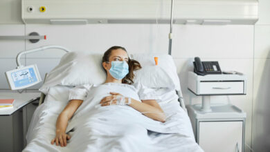 Sick female lying on bed in ICU. Mid adult patient is looking away while wearing protective face mask. She is in hospital during COVID-19 pandemic.