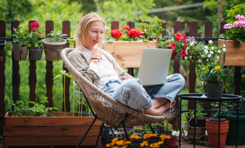 Beautiful blond middle-aged woman relaxing on the terrace of a country house in summer and using a laptop for online work balcony flower news