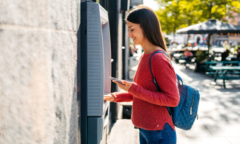 Young woman withdrawing money using phone at the ATM machine on the street in Copenhagen in Denmark.